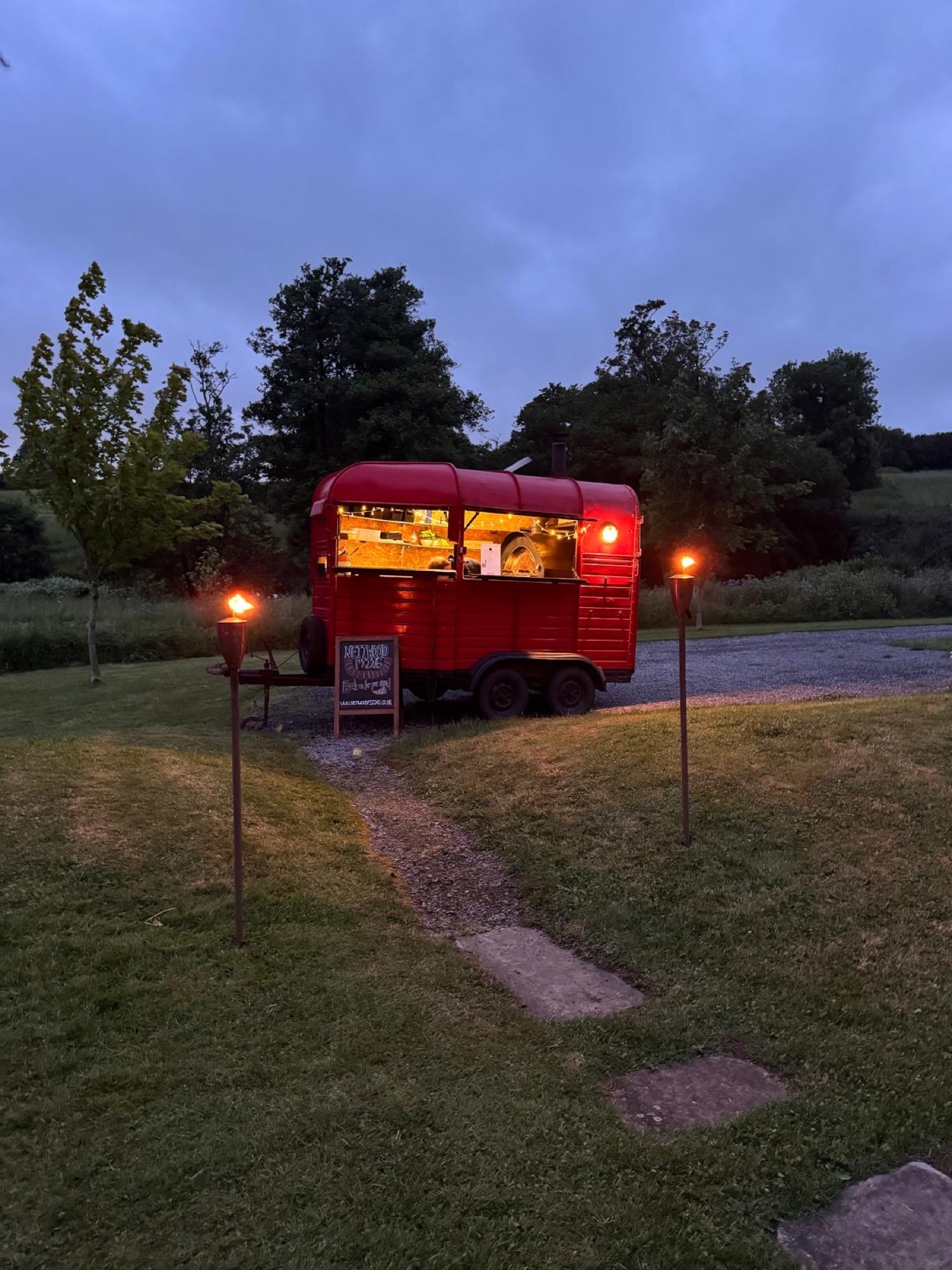 The red Nettwood horsebox at dusk, with a gravel path lit by two flaming torches